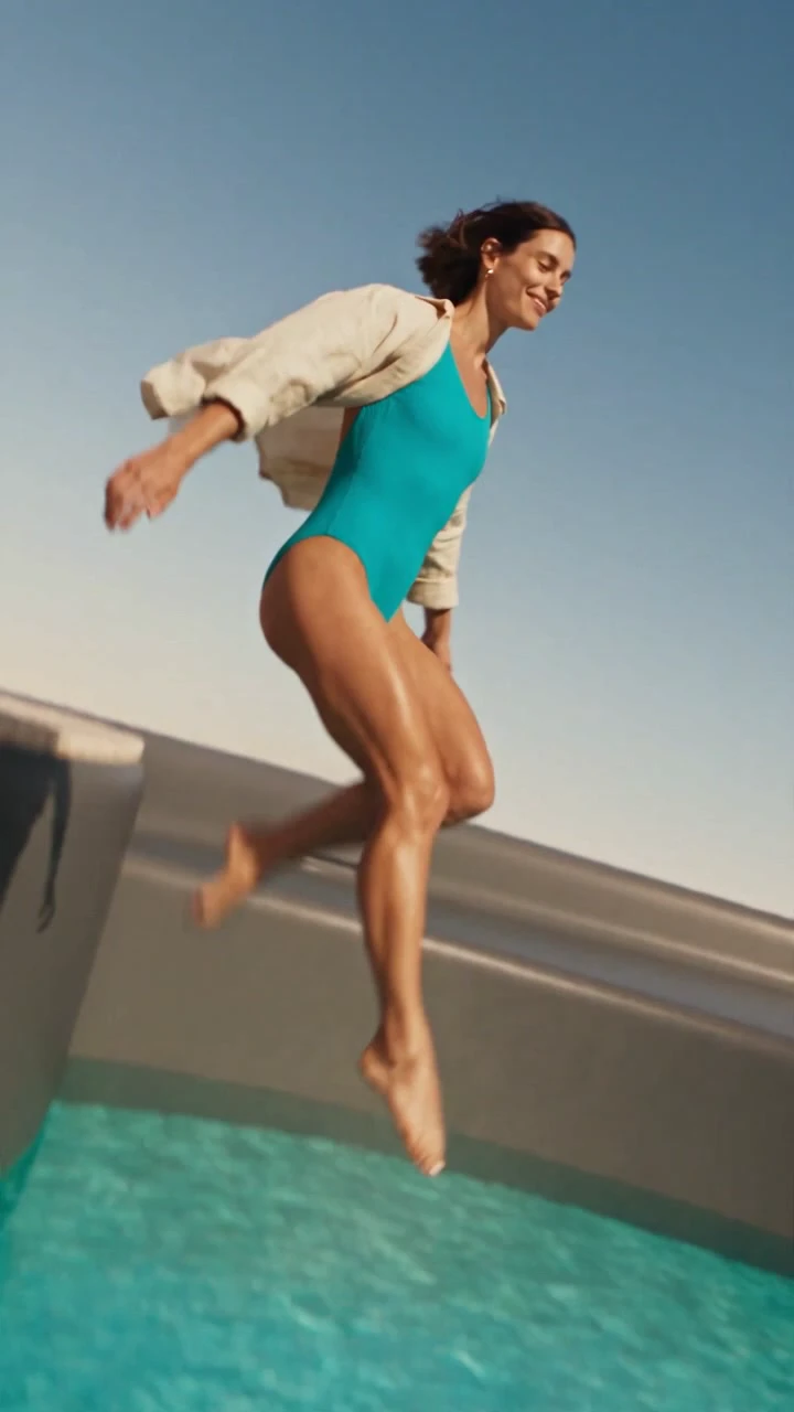 A woman jumping into a turquoise pool inside the lid of a giant olive travel cup.