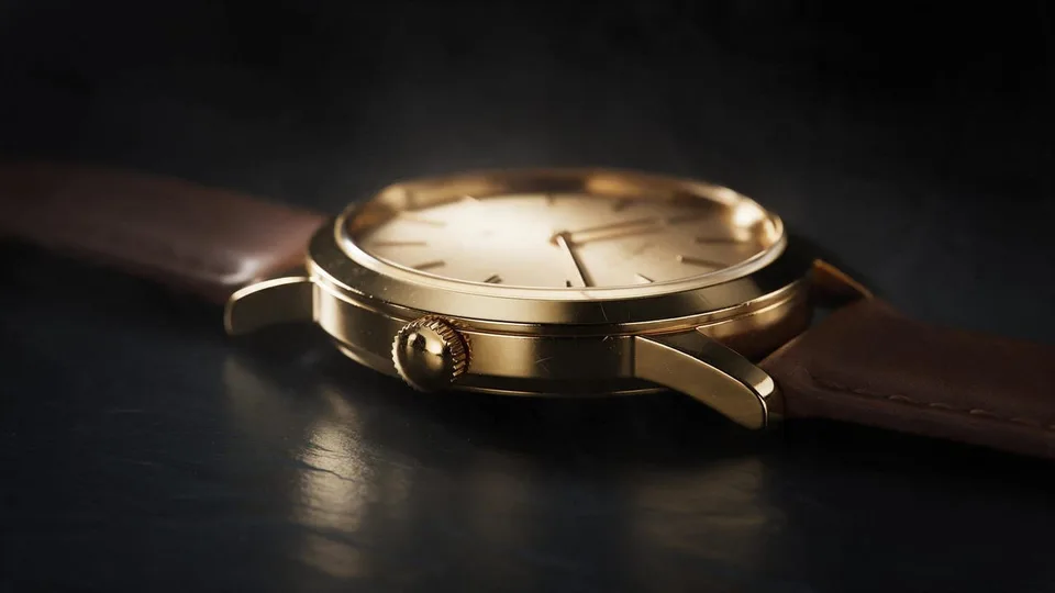 The knurled gold crown of the watch stands perfectly still on the case edge, the gold shimmering as light glides across it, the thin second hand sweeping on the champagne dial behind. No people and no hands in the shot.